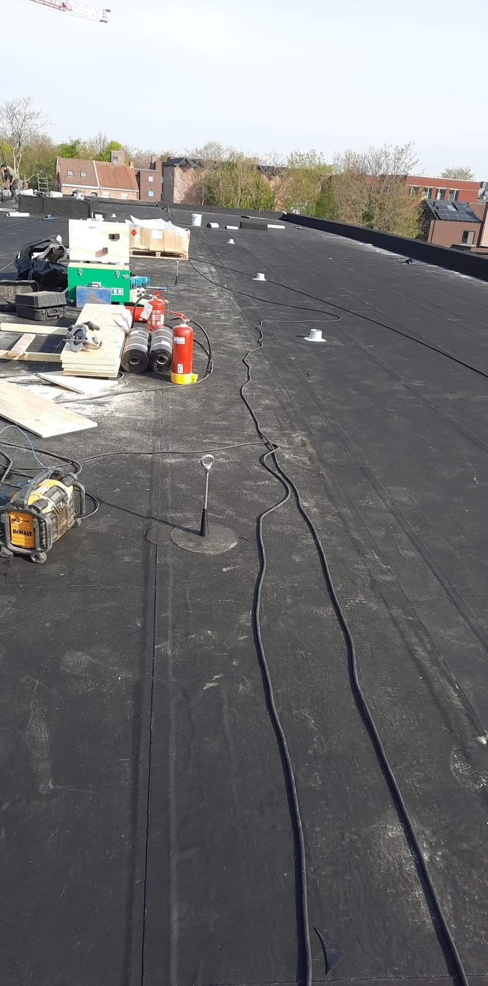 Czarny, płaski dach pokryty papą z widocznymi narzędziami, rolkami papy, butlami z gazem, oraz przewodami elektrycznymi podczas prac dekarskich w słoneczny dzień.