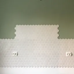 Białe, heksagonalne płytki częściowo ułożone na ścianie z zielonym fragmentem, widoczne dwa gniazdka elektryczne.