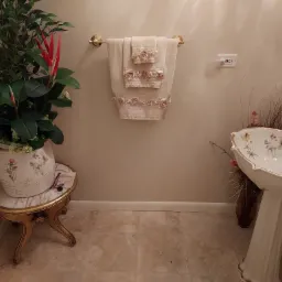 Umywalka kolumnowa w stylu vintage z dekoracyjnymi zdobieniami i złotą armaturą, obok wazon z suchymi trawami, na ścianie wieszak na ozdobne ręczniki, całość w ciepłej tonacji kolorystycznej.