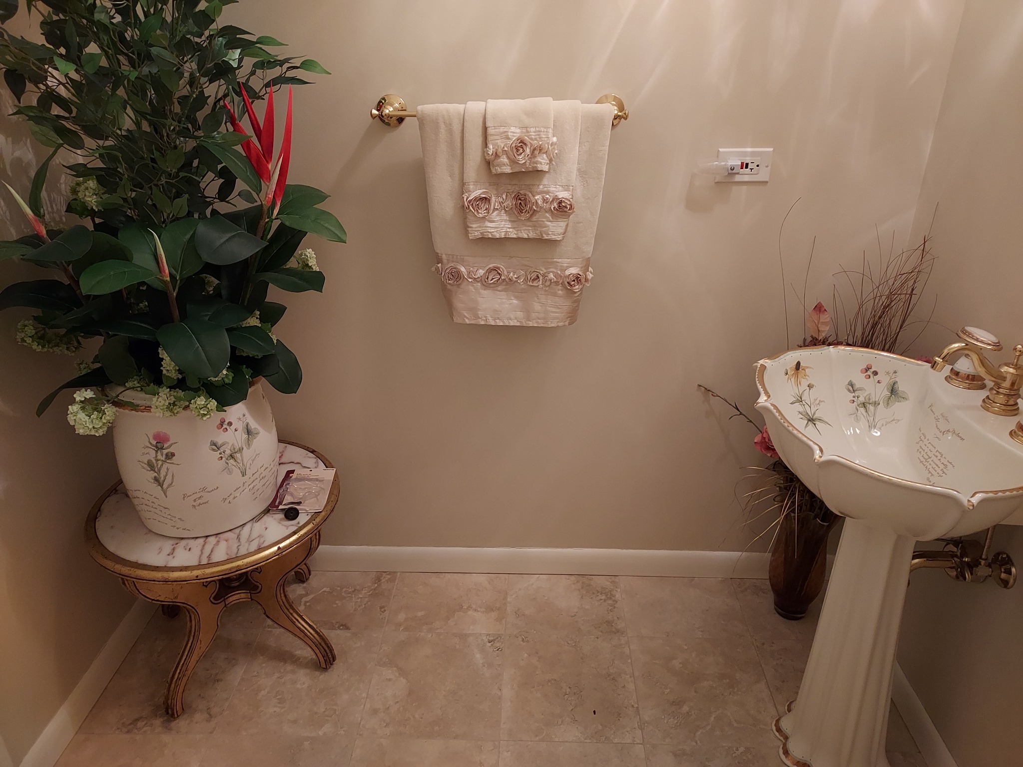 Umywalka kolumnowa w stylu vintage z dekoracyjnymi zdobieniami i złotą armaturą, obok wazon z suchymi trawami, na ścianie wieszak na ozdobne ręczniki, całość w ciepłej tonacji kolorystycznej.