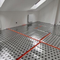 B.M-Bud Gorzów Wielkopolski - Przygotowanie do wylewki podłogowej w poddaszu: folia izolacyjna z nadrukiem ułożona na podłodze, wyznaczona pomarańczową taśmą, widoczny grzejnik i okno dachowe.