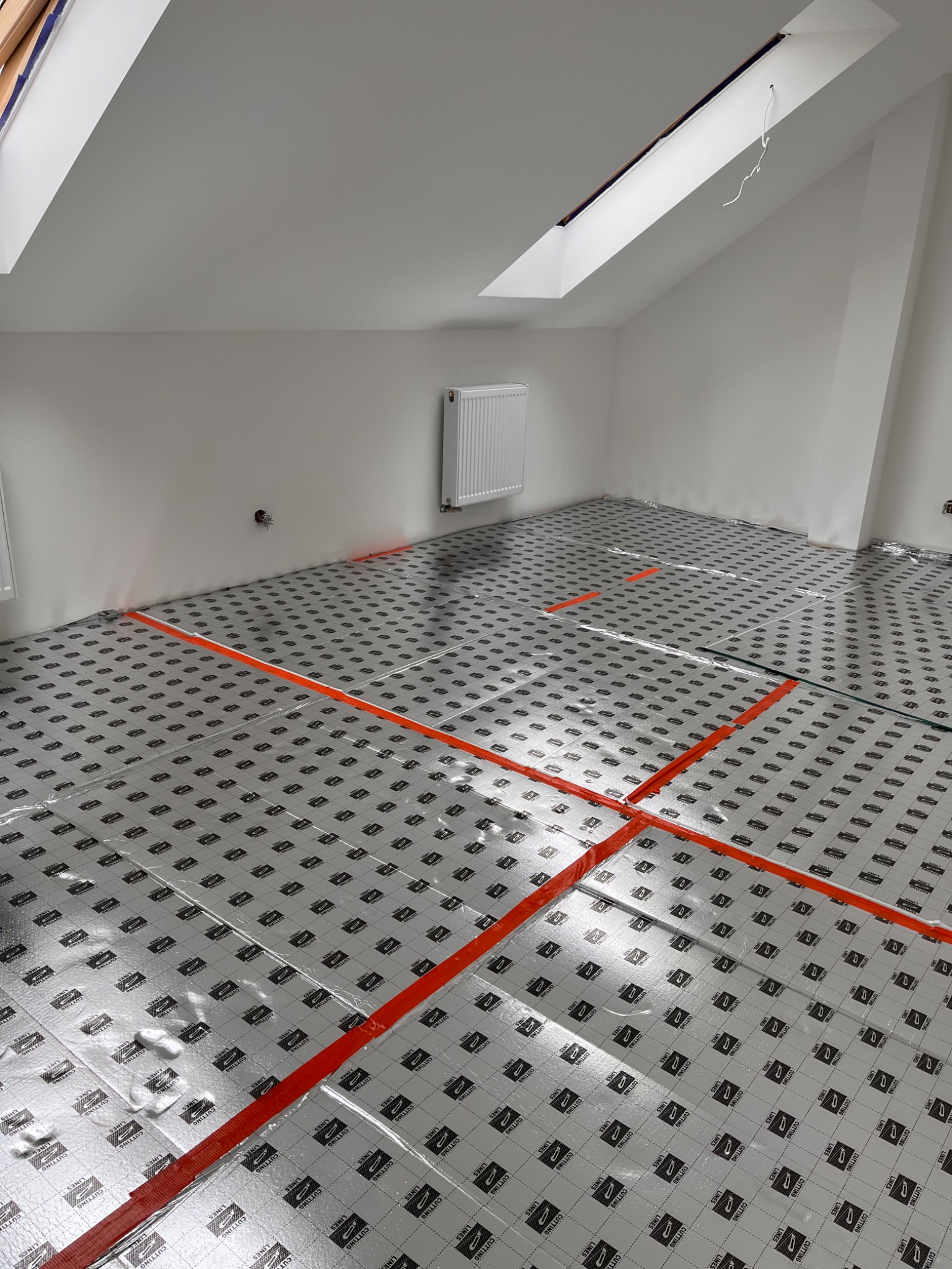 Przygotowanie do wylewki podłogowej w poddaszu: folia izolacyjna z nadrukiem ułożona na podłodze, wyznaczona pomarańczową taśmą, widoczny grzejnik i okno dachowe.