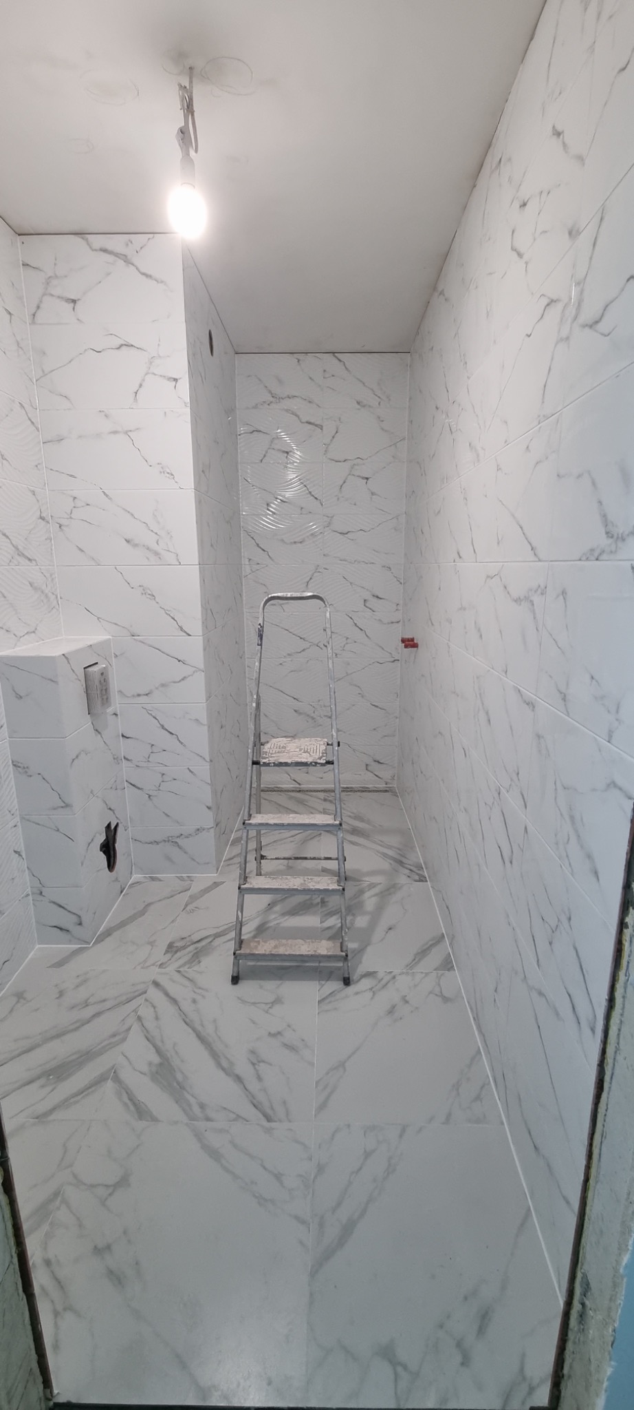 Wnętrze łazienki wykończone białymi płytkami imitującymi marmur, z widoczną drabinką malarską i wystającą żarówką.