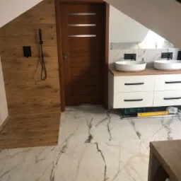 Wykończona łazienka na poddaszu z drewnopodobną kabiną prysznicową, marmurowymi płytkami podłogowymi i podwójną umywalką nablatową na białej szafce z czarnymi uchwytami.
