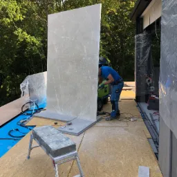 Pracownik w niebieskiej koszulce i spodniach, z zielonym wózkiem narzędziowym, szlifuje dużą, pionową płytę z szarego marmuru na tarasie w trakcie budowy, z widocznymi drzewami w tle.