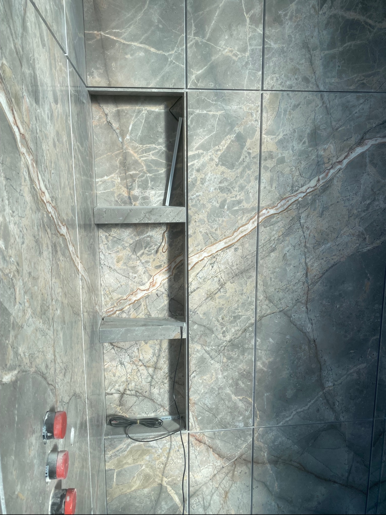 Ściana łazienki wyłożona szarymi płytkami imitującymi marmur, z wnęką z dwiema półkami i wystającymi przewodami elektrycznymi.
