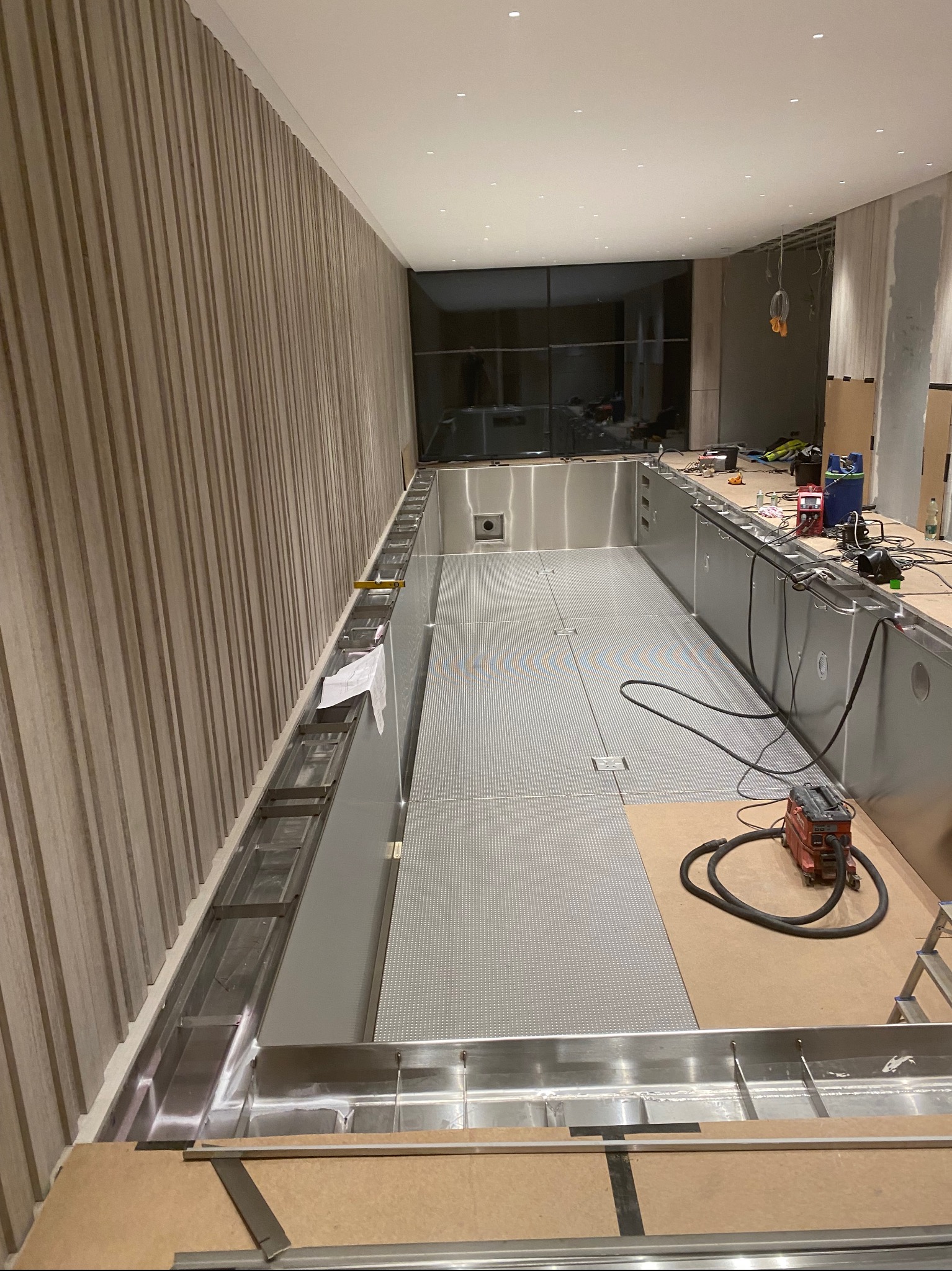 Wnętrze w trakcie budowy basenu ze stali nierdzewnej z drewnianą boazerią pionową na ścianie, widoczne narzędzia i materiały budowlane.