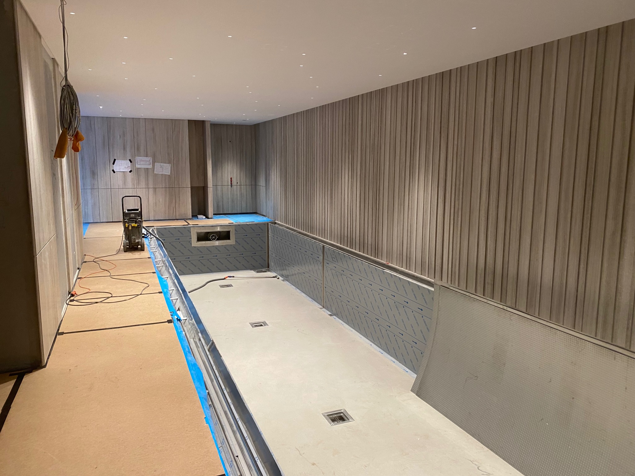 Wnętrze w trakcie budowy lub remontu, prawdopodobnie basenu wewnętrznego, z wykończonymi ścianami z paneli dekoracyjnych, przygotowaną podłogą i częściowo wyłożonymi ścianami niecką basenową,...