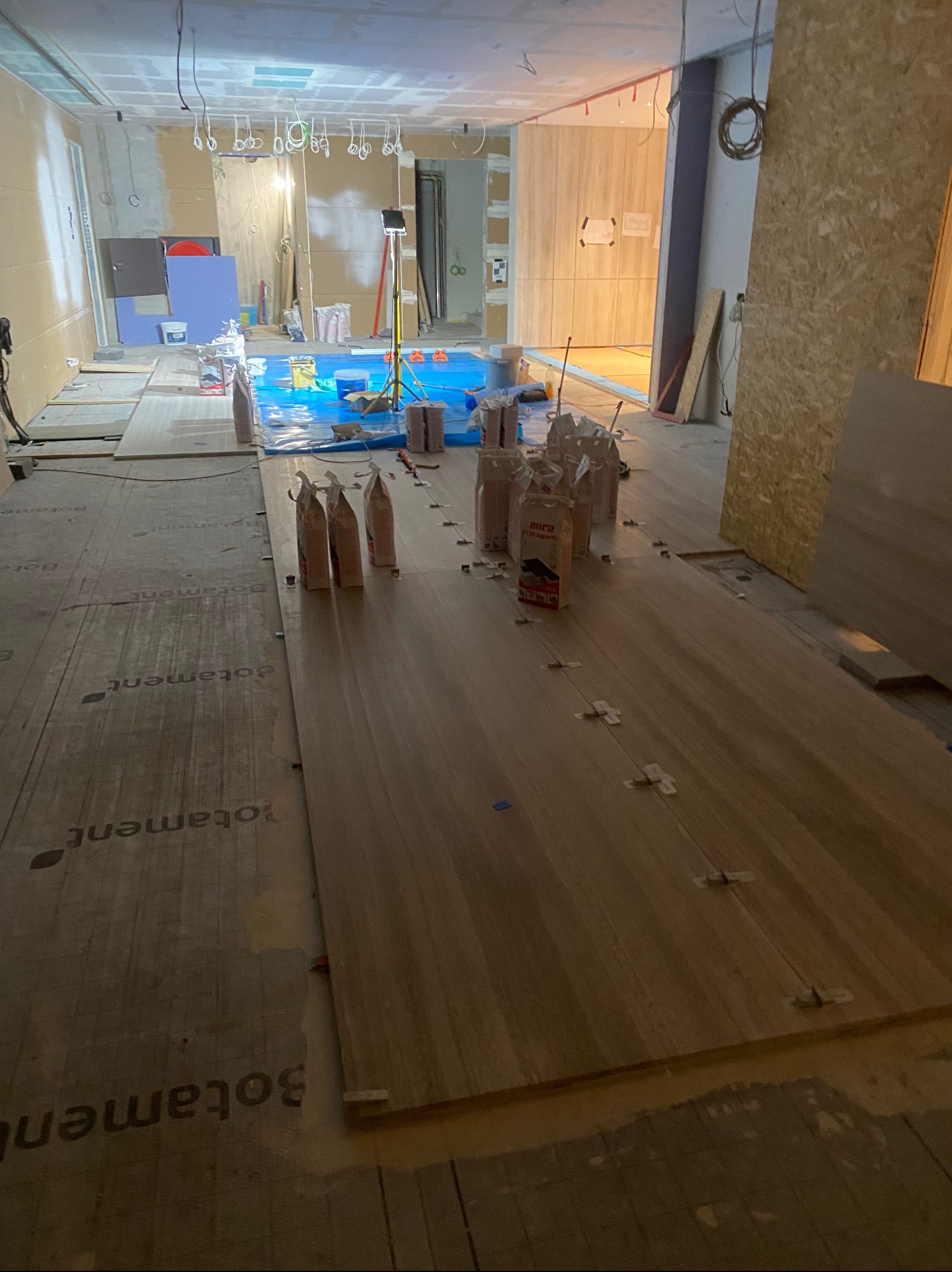 Wnętrze w trakcie remontu, widoczne płyty podłogowe z drewnopodobnym wzorem ułożone na podkładzie, paczki zaprawy klejowej MIRA i Botament, krzyżyki dystansowe, w tle zabezpieczona folią podłoga...
