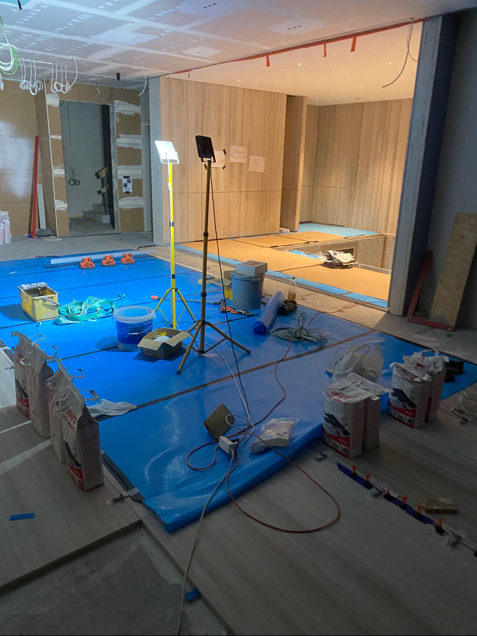 Wnętrze w trakcie układania płytek podłogowych, zabezpieczone niebieską folią ochronną, widoczne worki z zaprawą, poziomica laserowa na statywie, wiadro z klejem oraz listwy dystansowe.