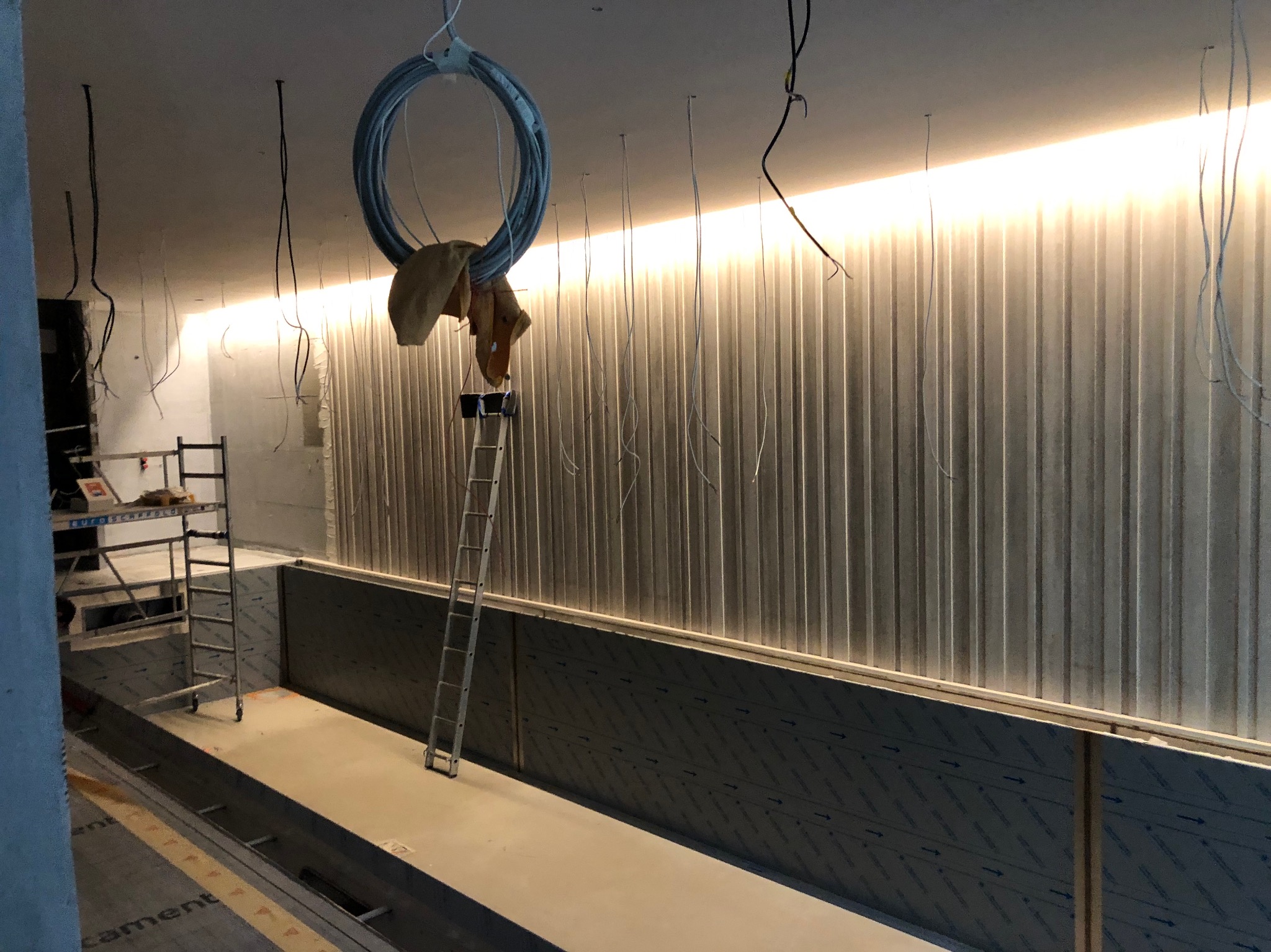 Wnętrze w trakcie remontu z metalową ścianą z oświetleniem LED, widoczne drabiny, zwisające kable i materiały budowlane.