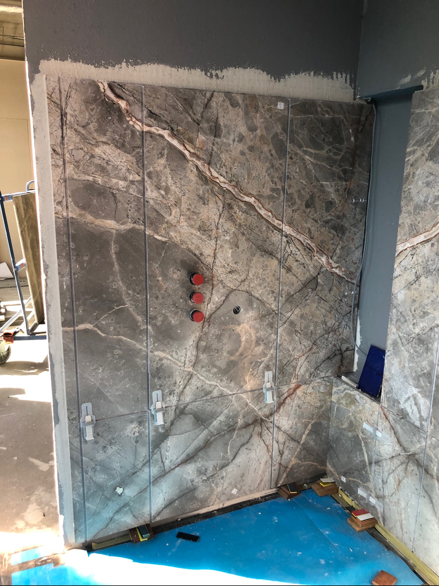 Montaż szarych płytek imitujących marmur w łazience, widoczne krzyżyki dystansowe, niebieska folia ochronna na podłodze i wystające czerwone zawory.