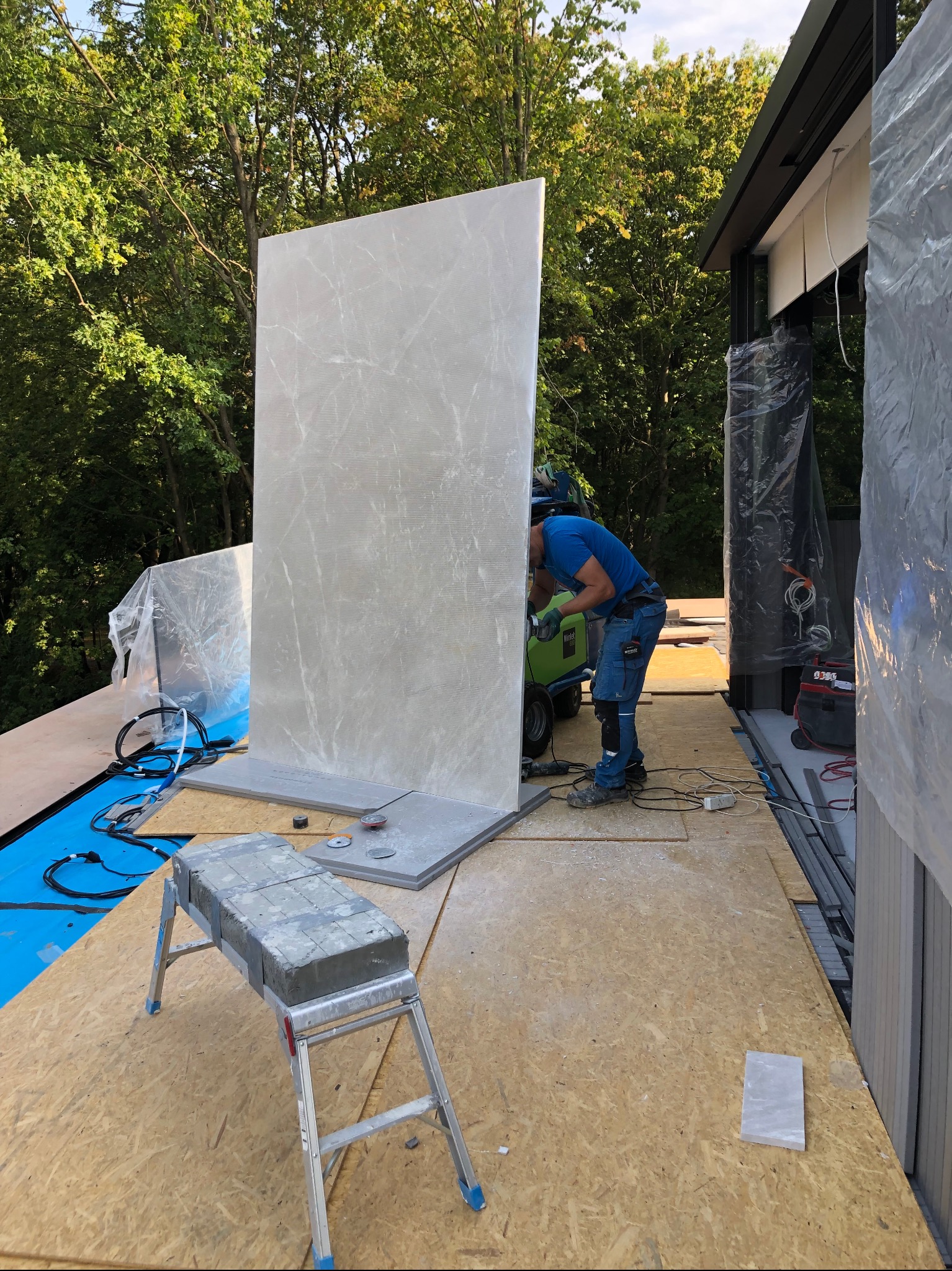 Pracownik w niebieskiej koszulce i spodniach, z zielonym wózkiem narzędziowym, szlifuje dużą, pionową płytę z szarego marmuru na tarasie w trakcie budowy, z widocznymi drzewami w tle.