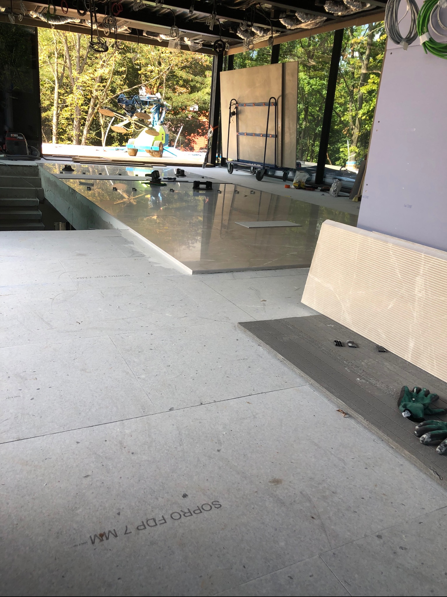 Wnętrze w trakcie prac wykończeniowych: przygotowanie podłoża, widoczne płyty podłogowe z napisem 'SOPRO FDP 7 MM', narzędzia, w tle maszyna do przenoszenia dużych płyt szklanych, widok na zewnątrz...