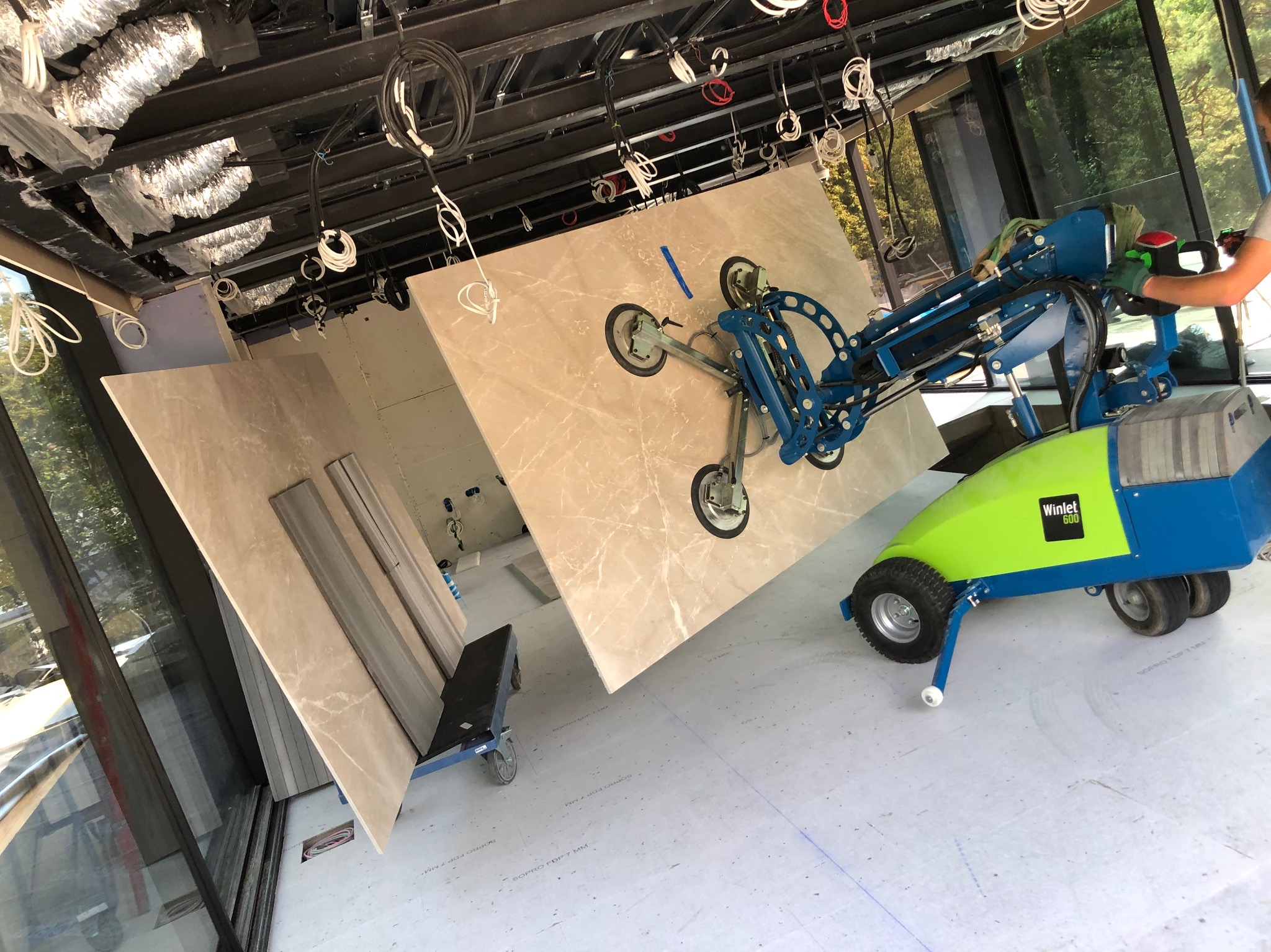 Montaż marmurowych płyt ściennych za pomocą specjalistycznego podnośnika Winlet 600 na budowie, widoczne przewody pod sufitem i duże okna.