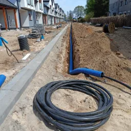Wykonanie wykopu pod kable elektryczne wzdłuż chodnika na tle szeregu nowych domów jednorodzinnych, widoczne bębny z kablami i niebieska rura karbowana w wykopie.