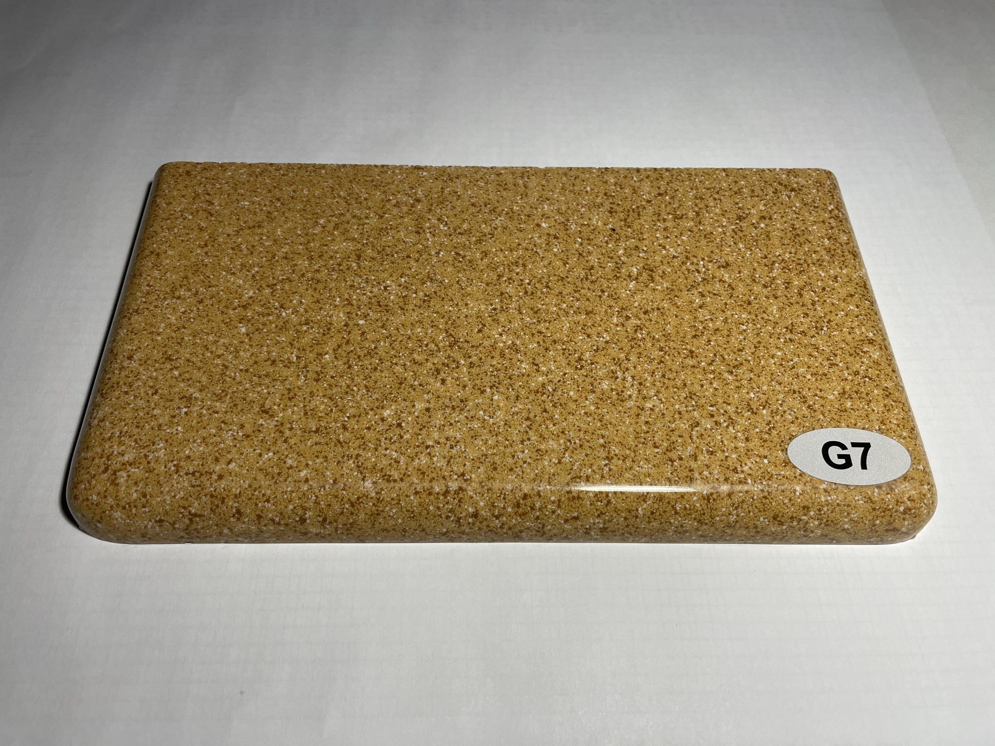 Próbka beżowego materiału kompozytowego z drobnym ziarnem i zaokrąglonymi krawędziami, oznaczona naklejką 'G7' w prawym dolnym rogu, leżąca na białym tle.