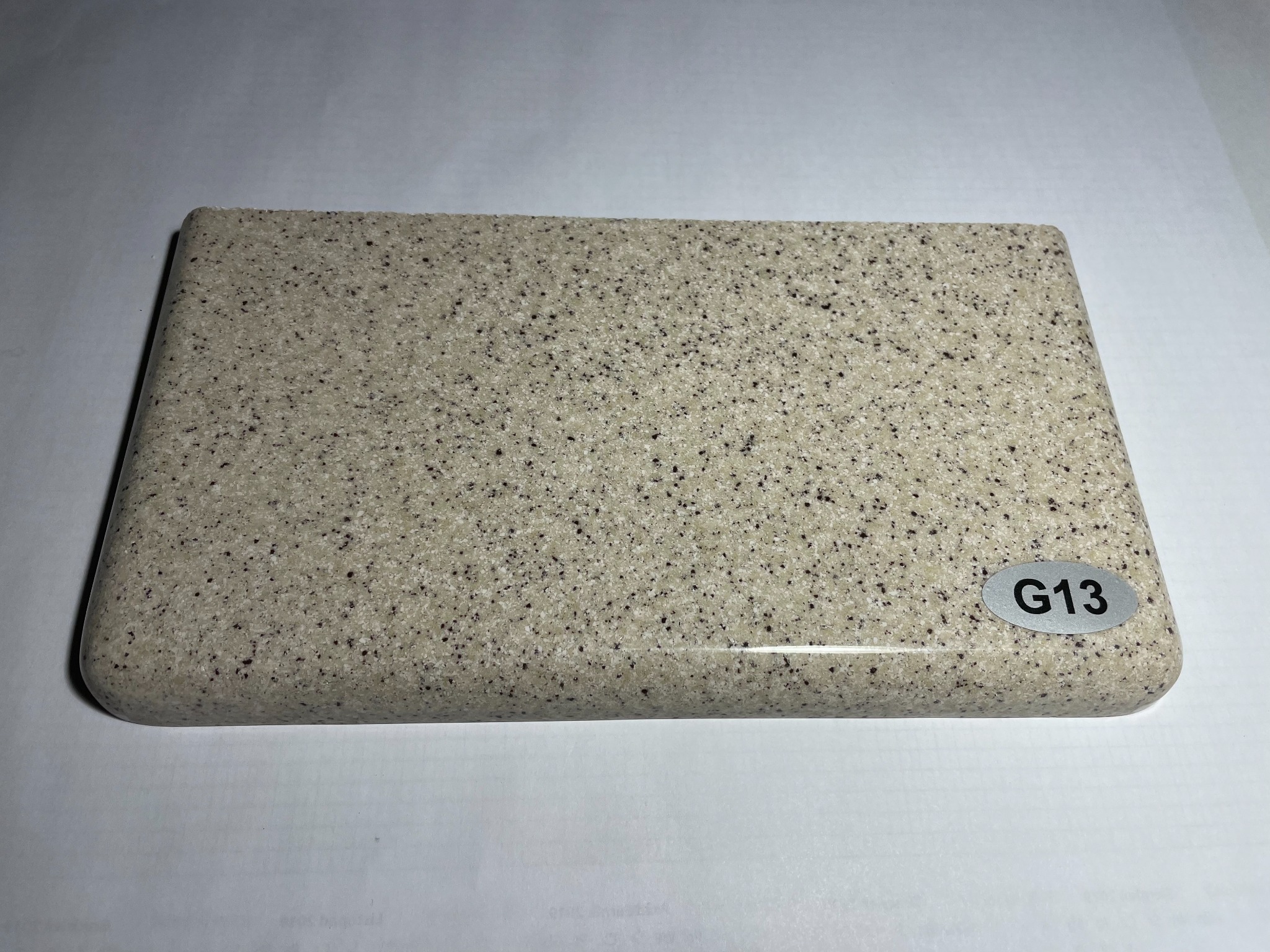 Próbka beżowego materiału kompozytowego z drobnymi, ciemnymi plamkami, o zaokrąglonych krawędziach, oznaczona naklejką z symbolem 'G13'.