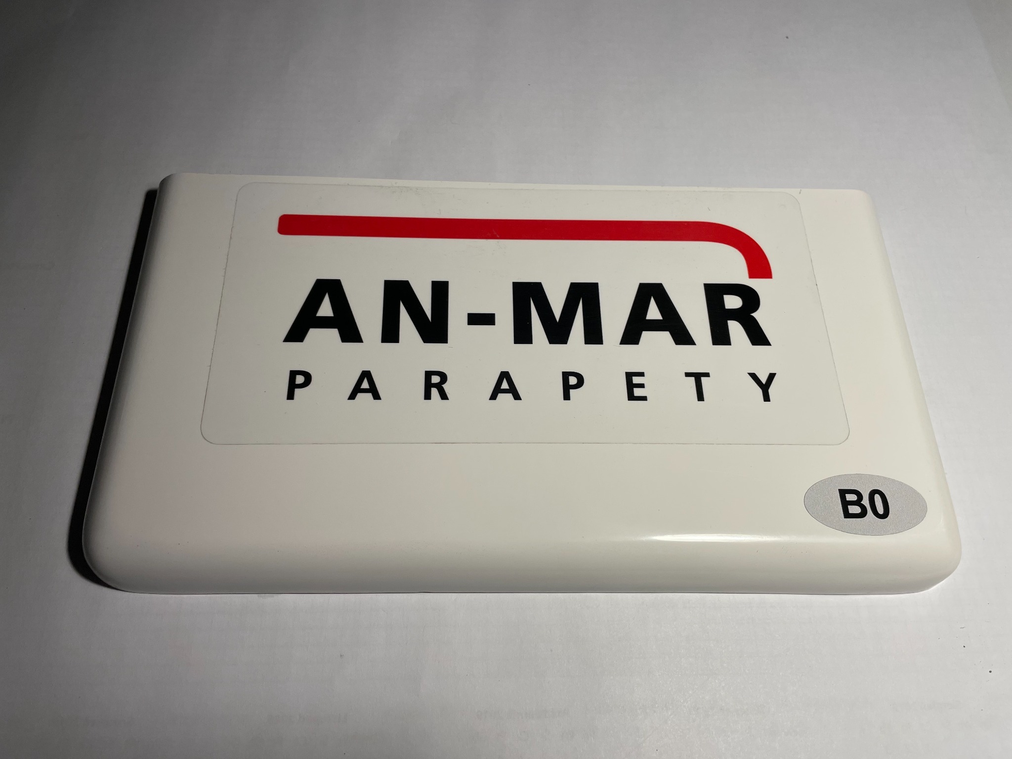 Biała próbka z nadrukowanym logo firmy 'AN-MAR PARAPETY' z czerwoną linią nad nazwą i oznaczeniem 'B0' w owalnej naklejce w prawym dolnym rogu, leżąca na białym tle.