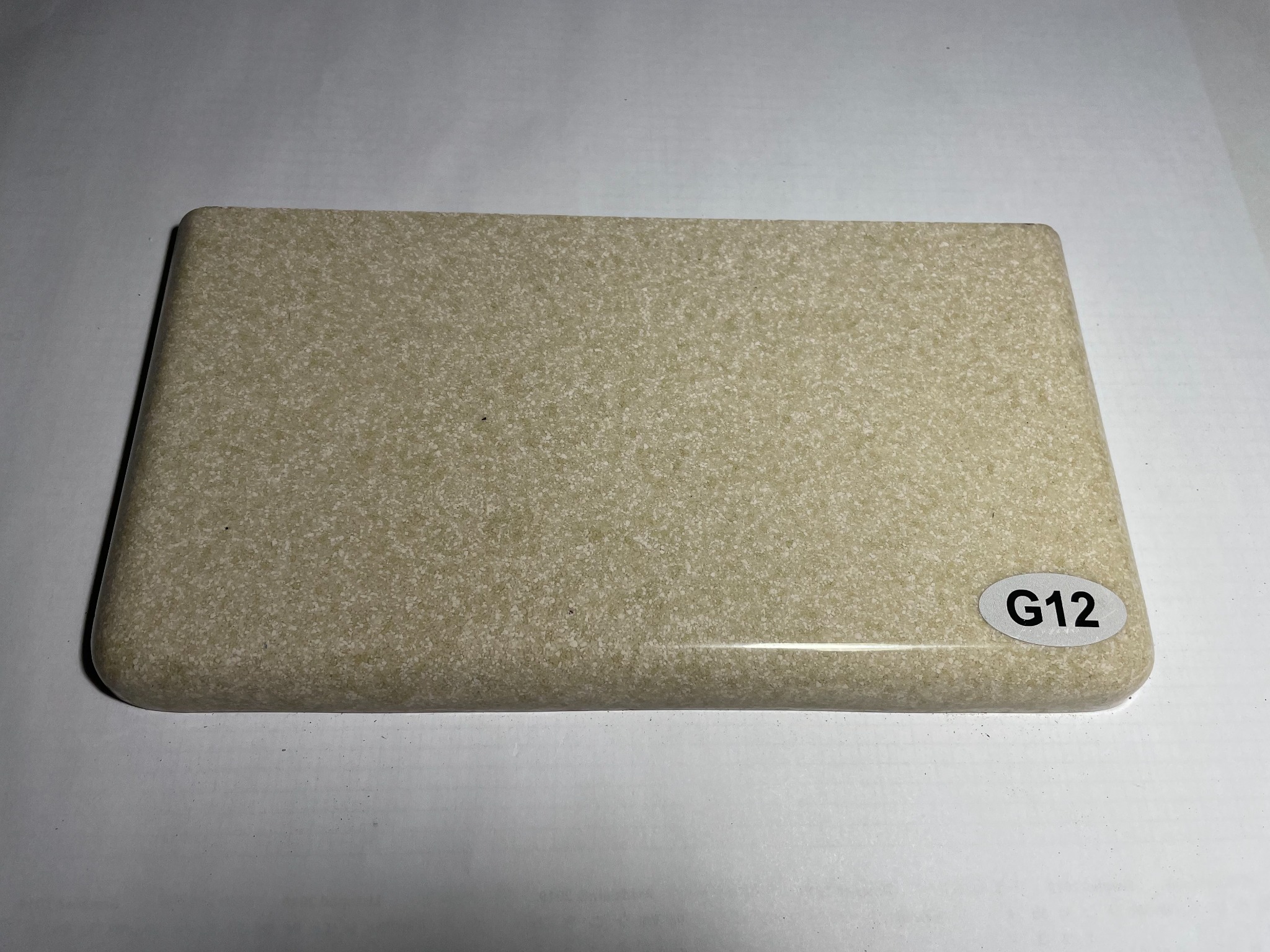 Próbka beżowego materiału kompozytowego o fakturze drobnego kamienia, z zaokrąglonym brzegiem i naklejką z oznaczeniem 'G12', leżąca na białym tle.