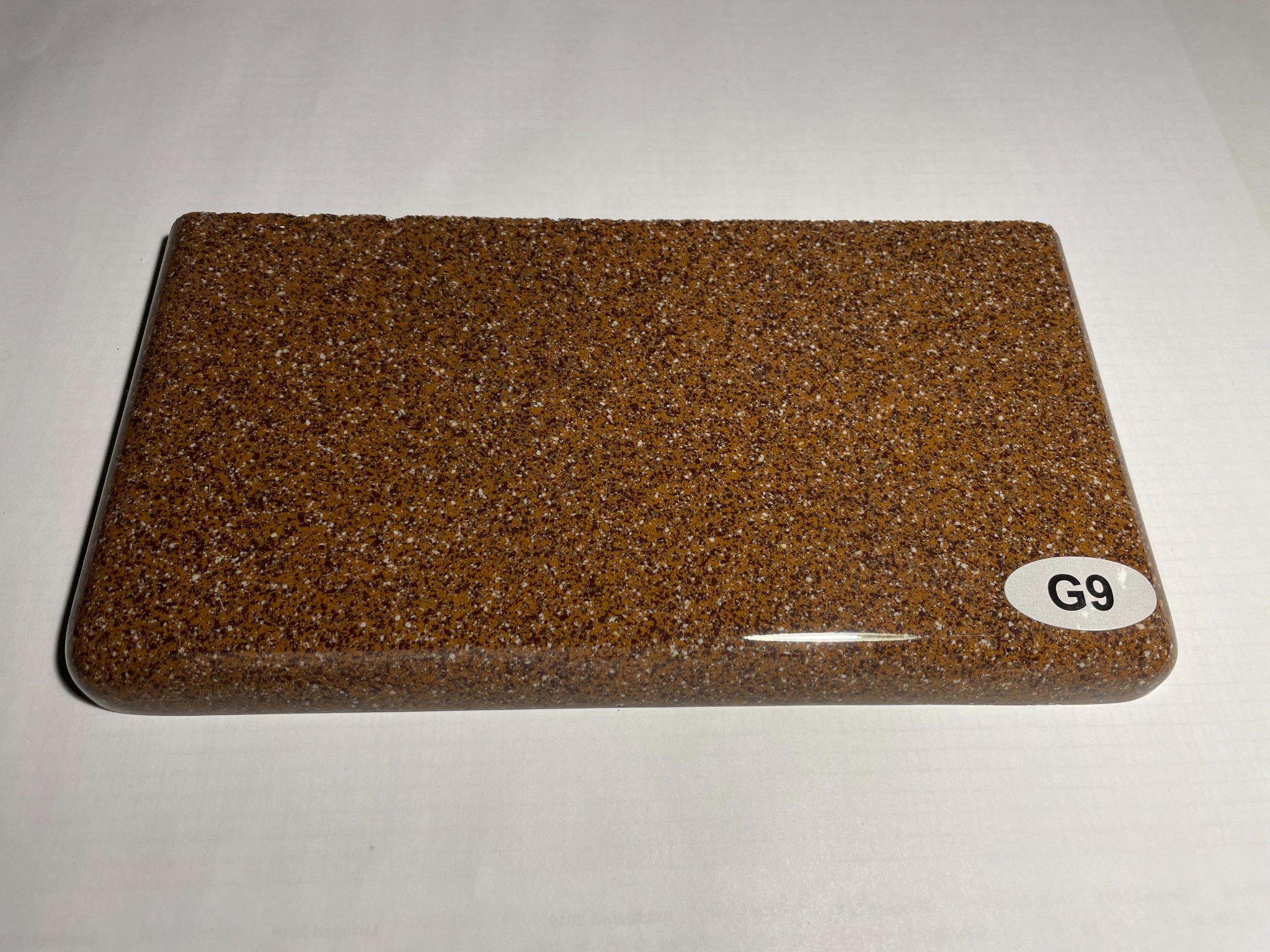 Próbka brązowego parapetu z konglomeratu z białymi i ciemnobrązowymi drobinami, oznaczona naklejką 'G9', leżąca na białym tle.