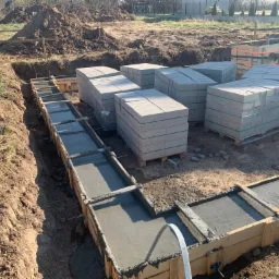 Wylany beton w szalunku fundamentowym, obok ułożone palety z bloczkami betonowymi, widok na wykop i pryzmę ziemi w tle.