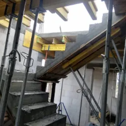 Wylewanie betonowych schodów w surowym stanie z widocznym szalunkiem i podporami stalowymi w budynku z białych bloczków.