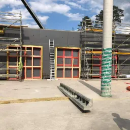 Budowa domu: szalunki okienne z czerwonymi ramami, rusztowania, betonowa podłoga i filar zabezpieczony folią, widok na plac budowy w słoneczny dzień.