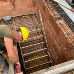 Murarz w żółtym kasku pracuje nad wznoszeniem ściany z czerwonej cegły dziurawki, widoczne zbrojenie i szalunek, poziomica, wiadro i drewniane schody do wykopu.