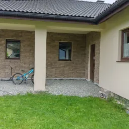 Wykończona ściana frontowa domu z kamienia ozdobnego, widoczny fragment dachu z brązową dachówką, okna z brązowymi ramami, kolumna podtrzymująca zadaszenie oraz rower i hulajnoga zaparkowane obok.