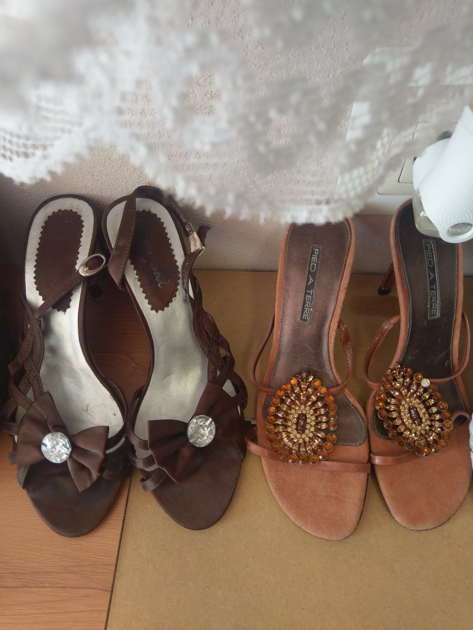 Dwie pary damskich sandałów na obcasie: brązowe z kokardkami i kryształkami oraz pomarańczowe zdobione bursztynowymi aplikacjami w stylu boho, ustawione obok siebie na drewnianej powierzchni,...