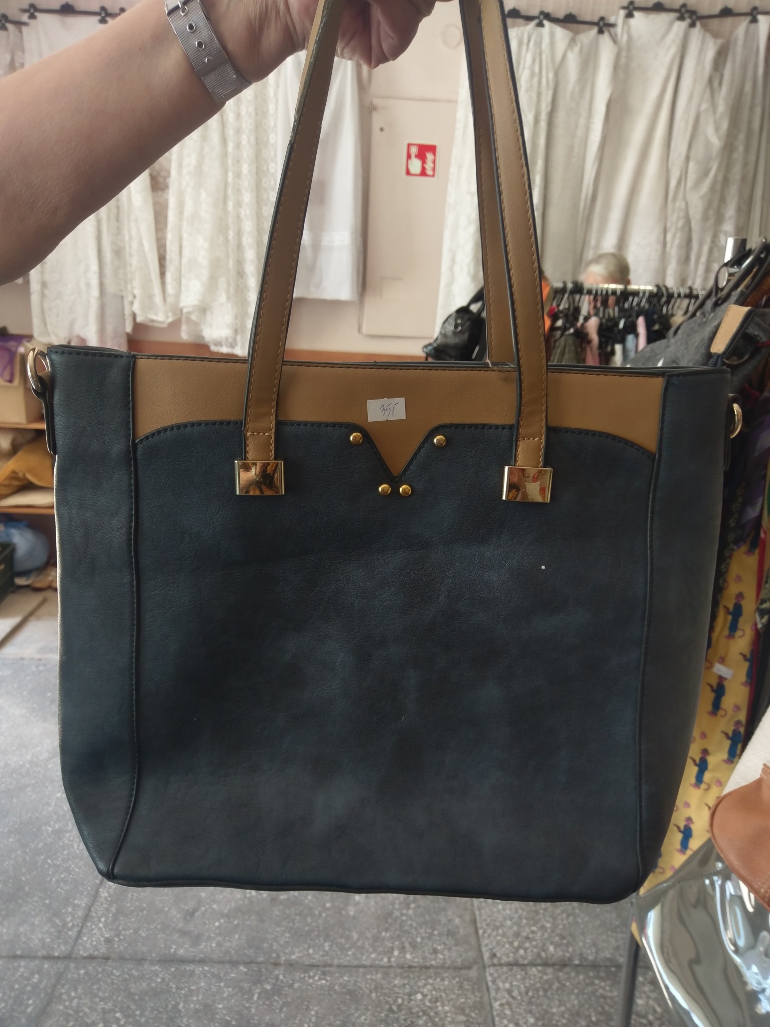 Czarno-brązowa torebka damska z krótkimi uchwytami, prezentowana na tle wieszaków z ubraniami i białych zasłon.