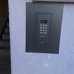 Zamontowany, nowoczesny domofon ACO z klawiaturą numeryczną na fioletowej ścianie budynku, obok okna. Widoczne schody wejściowe.