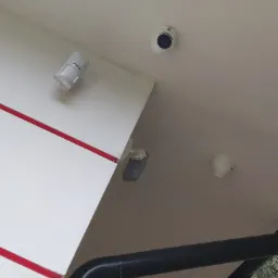 Widok z dołu na sufit z zamontowanymi elementami systemu alarmowego: okrągła kamera, czujnik ruchu i czujnik dymu, z widoczną czerwoną listwą dekoracyjną na ścianie i fragmentem czarnej rury...