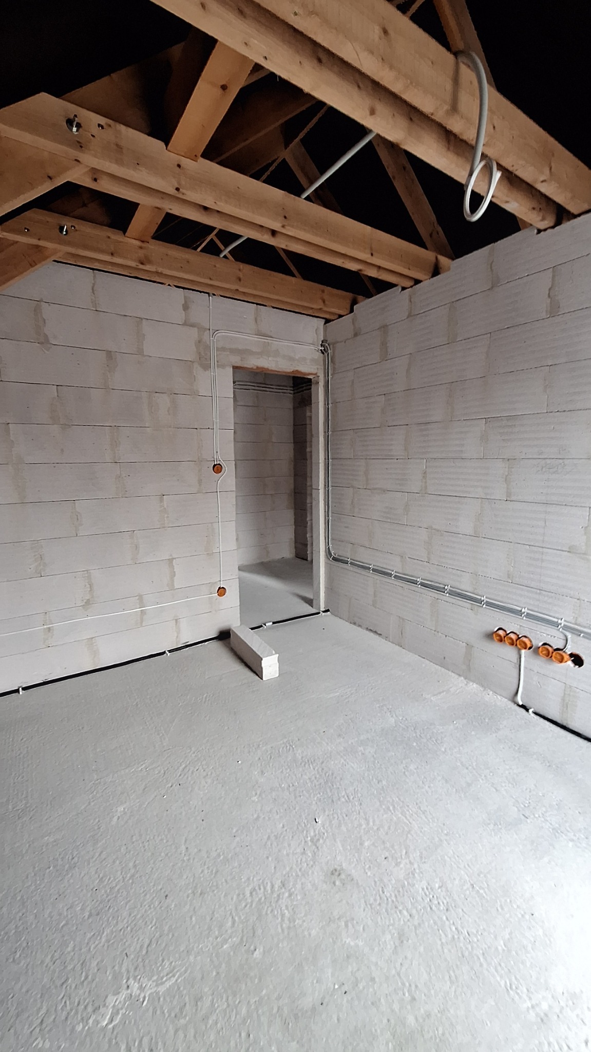 Surowe wnętrze z bloczków betonowych i drewnianą konstrukcją dachu, widoczne przewody elektryczne w peszlach i puszki instalacyjne, surowy betonowy podkład podłogowy.