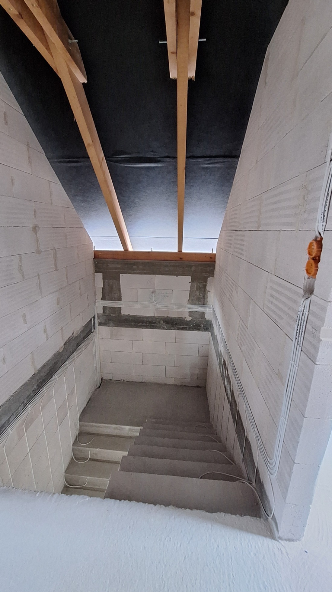Surowe wnętrze w trakcie budowy: schody betonowe, ściany z białych bloczków, widoczne kable elektryczne w peszlach, drewniana konstrukcja dachu.