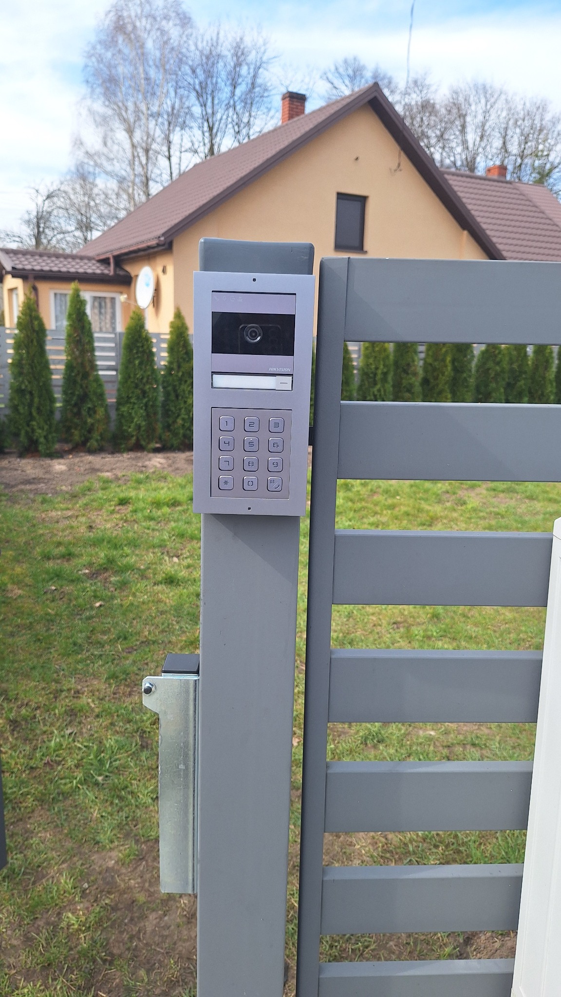 Szary wideofon Hikvision z klawiaturą numeryczną zamontowany na słupku ogrodzenia posesji. W tle dom jednorodzinny i tuje.