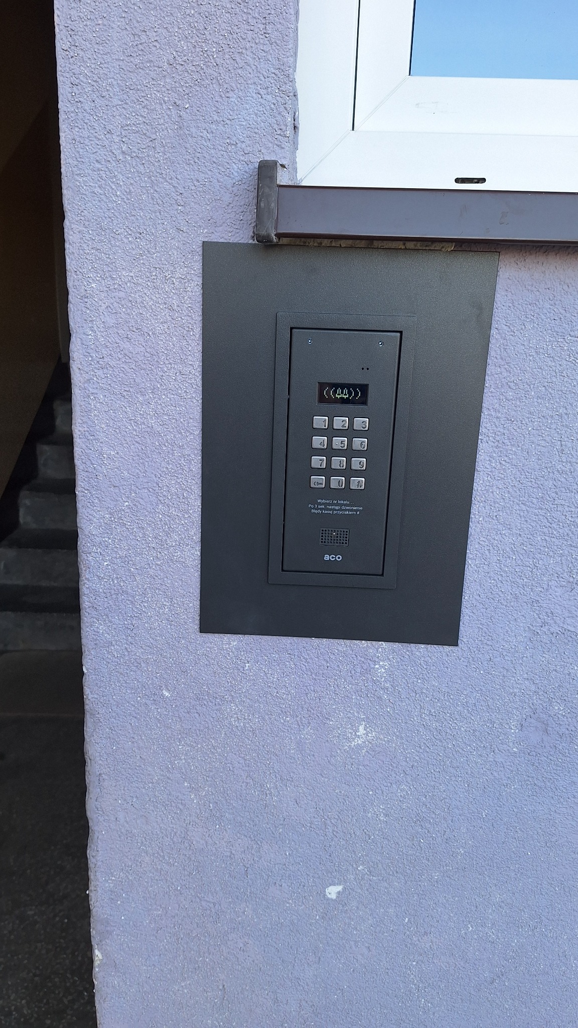 Zamontowany, nowoczesny domofon ACO z klawiaturą numeryczną na fioletowej ścianie budynku, obok okna. Widoczne schody wejściowe.