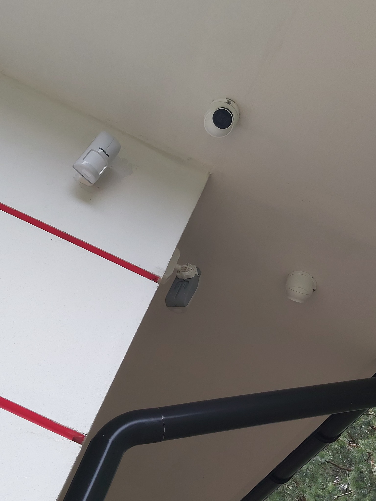 Widok z dołu na sufit z zamontowanymi elementami systemu alarmowego: okrągła kamera, czujnik ruchu i czujnik dymu, z widoczną czerwoną listwą dekoracyjną na ścianie i fragmentem czarnej rury...