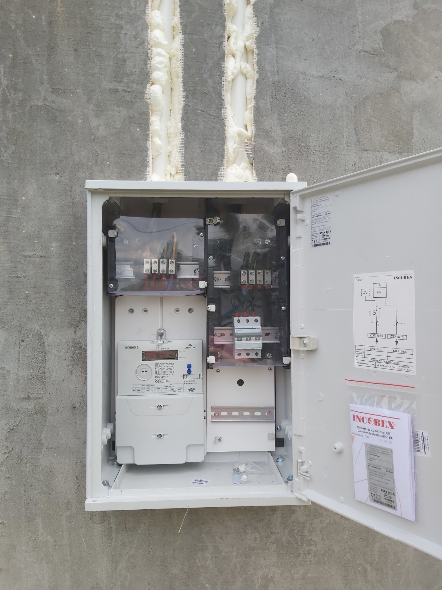 Otwarta skrzynka elektryczna z licznikiem i schematem instalacji na szarym, surowym tynku, widoczne dwie pionowe szczeliny wypełnione pianką montażową.
