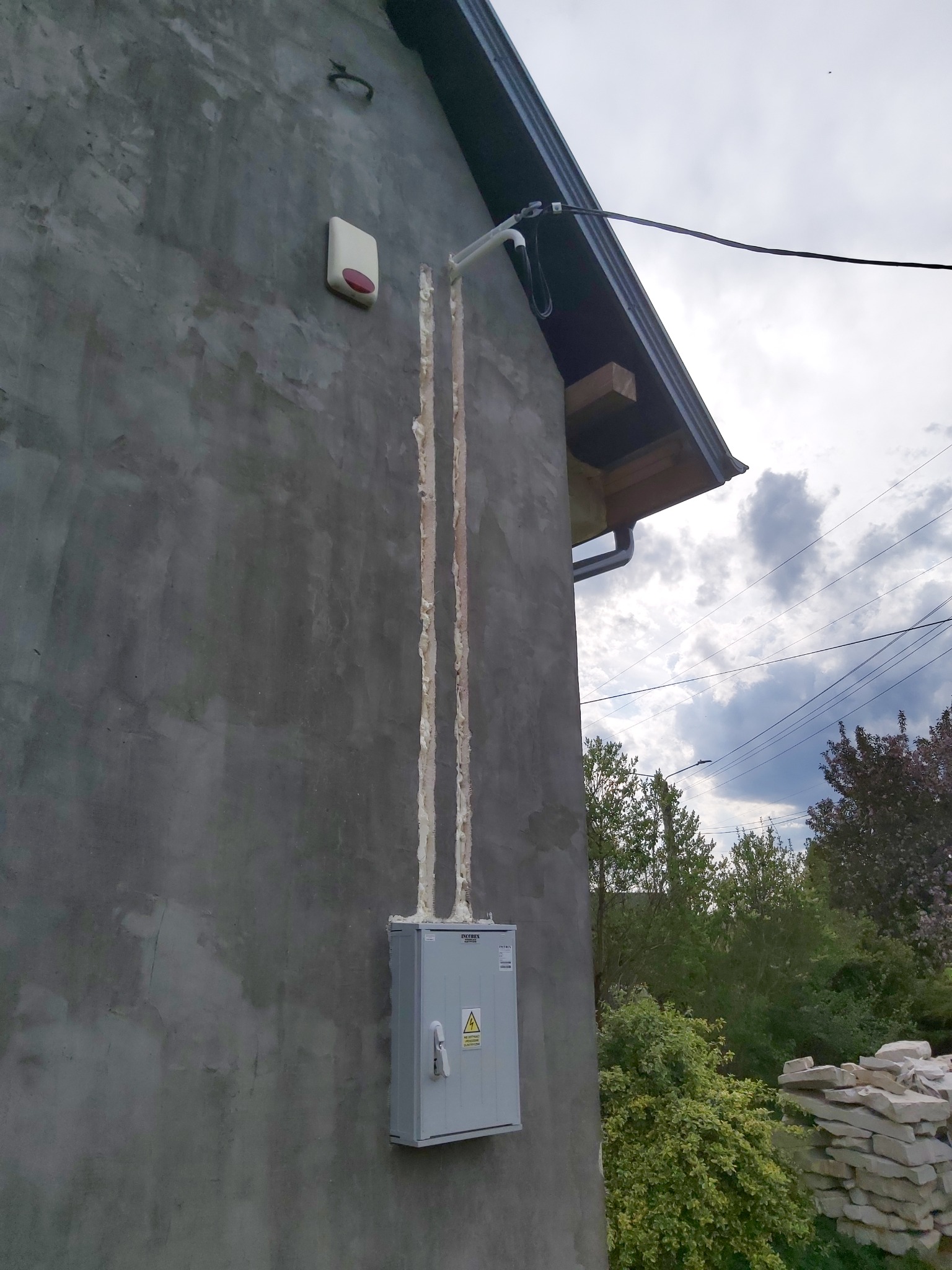 Szara skrzynka elektryczna z ostrzeżeniem o napięciu, zamontowana na surowej, szarej ścianie budynku, z widocznymi dwoma pionowymi rurami zabezpieczonymi pianką montażową, alarm zewnętrzny...