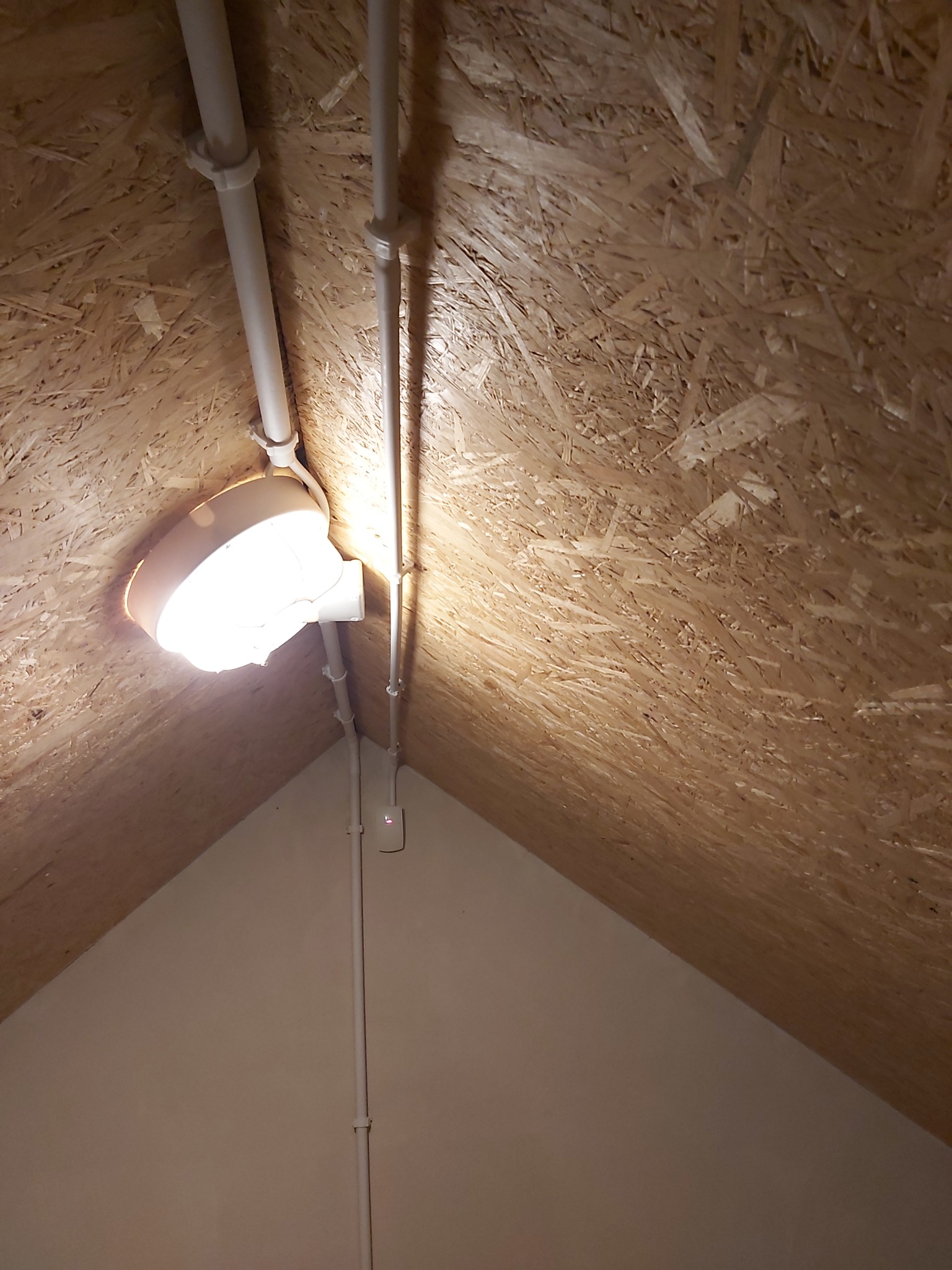 Białe rury instalacji elektrycznej przymocowane do drewnianej ściany z płyty OSB, oświetlone przez zamontowaną lampę sufitową.