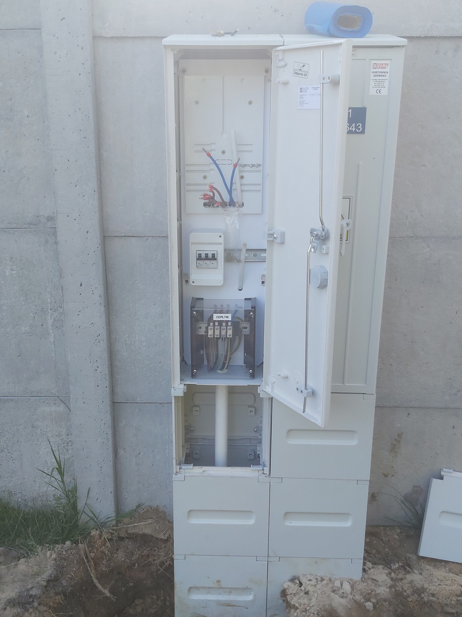 Otwarta szafka elektryczna z widocznymi przewodami, bezpiecznikami i złączami, umieszczona na zewnątrz budynku przy betonowym ogrodzeniu. Częściowo zakopana w ziemi. Na szafce widoczne naklejki...