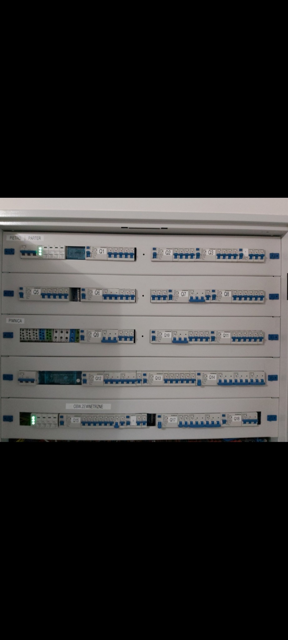 Rozdzielnica elektryczna z bezpiecznikami oznaczonymi Q1-Q18, z podziałem na obwody: Piętro/Parter, Piwnica i Obwód Zewnętrzny, niektóre wyłączniki w pozycji włączonej.