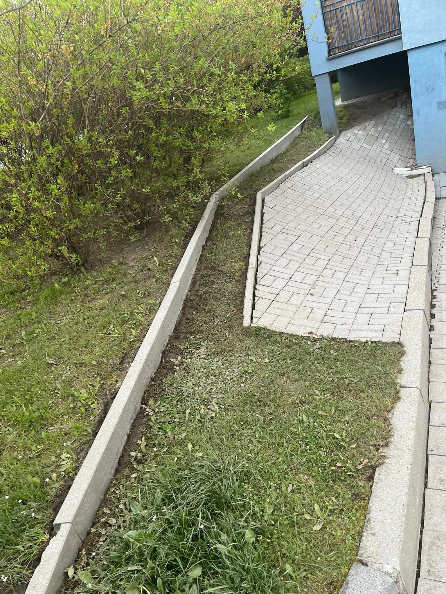 Ułożona kostka brukowa na podjeździe z widocznymi obrzeżami oraz fragmentem trawnika i krzewów w tle, z lekkim nachyleniem terenu.