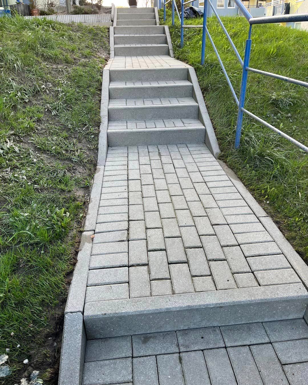Schody i ścieżka ułożone z szarej kostki brukowej prowadzące w górę zbocza porośniętego trawą, zabezpieczone niebieską barierką.