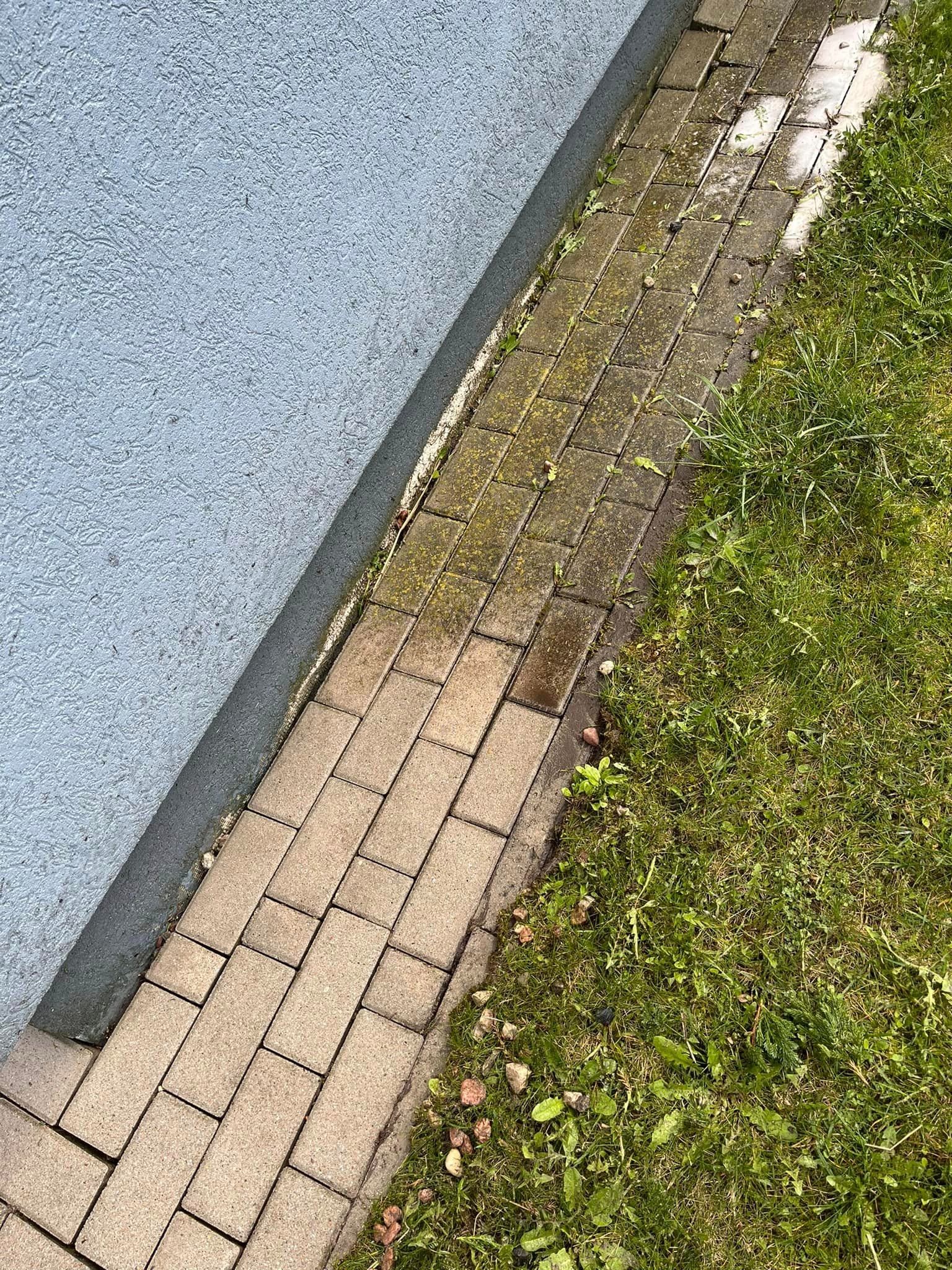 Częściowo wyczyszczona kostka brukowa przylegająca do ściany budynku, widoczny kontrast między czystą a zabrudzoną powierzchnią, obok rosnąca trawa i chwasty.