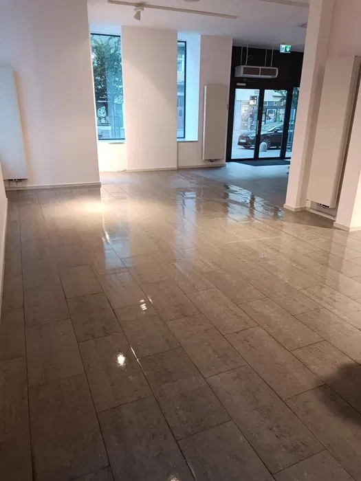 Świeżo umyta, lśniąca podłoga z płytek w pustym biurze, widoczne odbicia światła, białe ściany i przeszklone drzwi wejściowe.