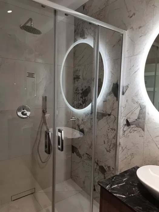 Nowoczesna kabina prysznicowa z przesuwnymi drzwiami, wykończona marmurowymi płytkami, z okrągłym lustrem z podświetleniem LED i armaturą w kolorze chromu.