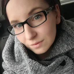 Portret kobiety w okularach i szarym, dzierganym swetrze z dużym kołnierzem, czarne oprawki okularów z widocznym logo na zauszniku.